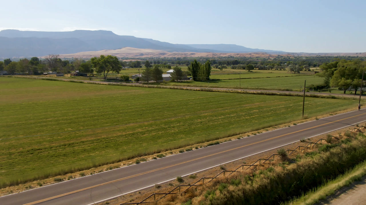 Palisades, Colorado farmland seen from above with a diagonal road across the lower frame, a utility pole near the roadside, tree clusters, small buildings, and hazy mountains in back.