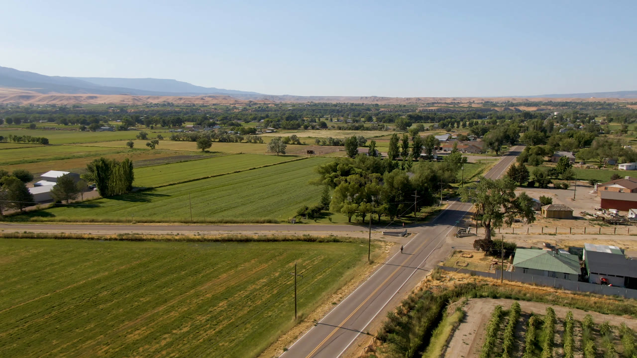 Wide aerial frame over Palisades farmland one second in, showing broad green fields, a road running through the lower frame, farm structures, tree rows, distant hills, clear sky, and no visible people.