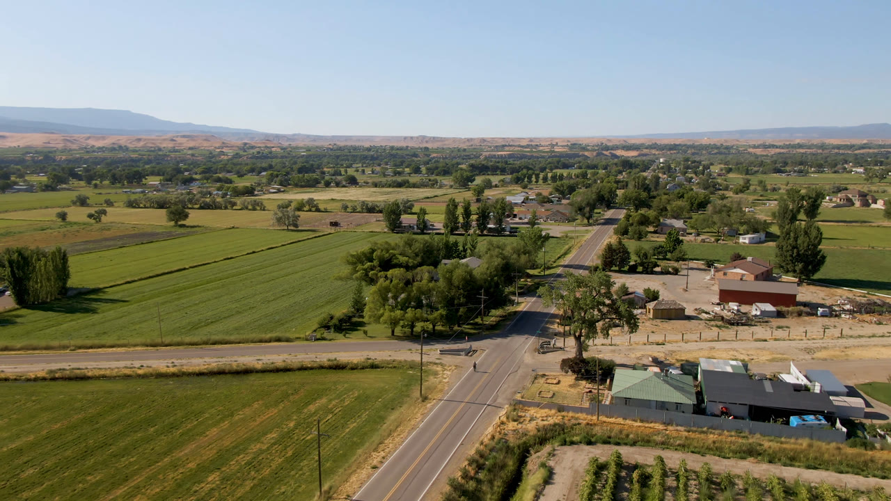 Wide aerial view of green fields, a road, farm buildings, trees, and distant hills.