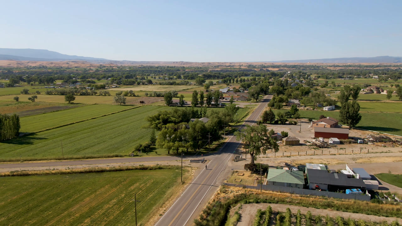 Second aerial frame in Palisades, Colorado showing the road more centrally, with farm buildings below, rectangular green fields, tree clusters, distant hills, and no visible people.