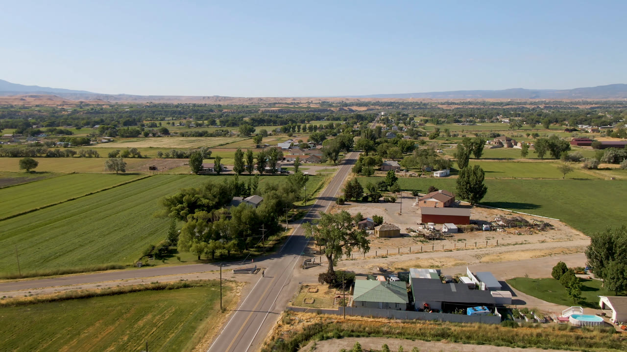 Wide aerial frame over Palisades, Colorado showing green farm fields, a paved road, clustered rural buildings, trees, distant hills, and no visible people.