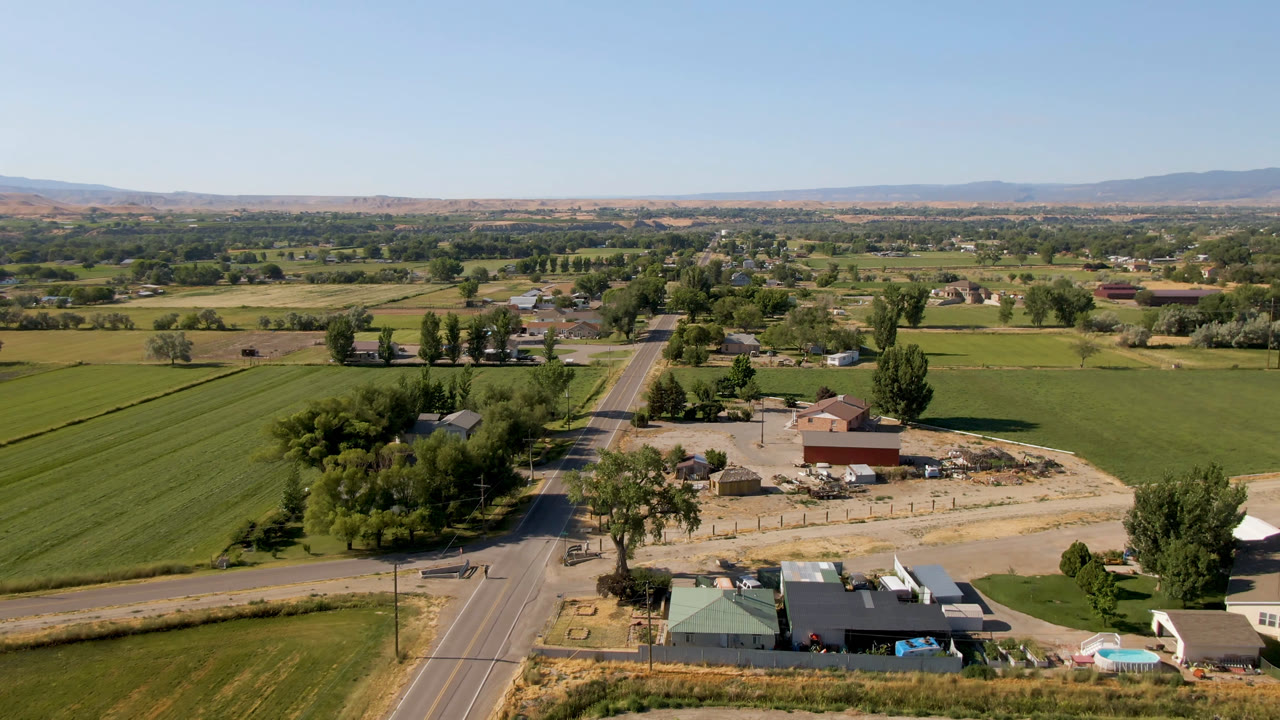 High aerial over a road, farm lots, green fields, tree clusters, and distant mountains.