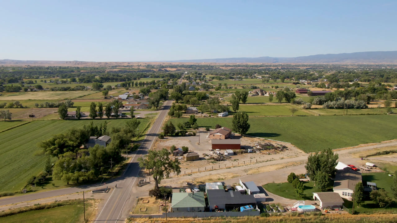 Aerial frame over Palisades, Colorado showing green fields, a paved road, scattered farm buildings, trees, dry foothills, distant mountains, and no visible people.