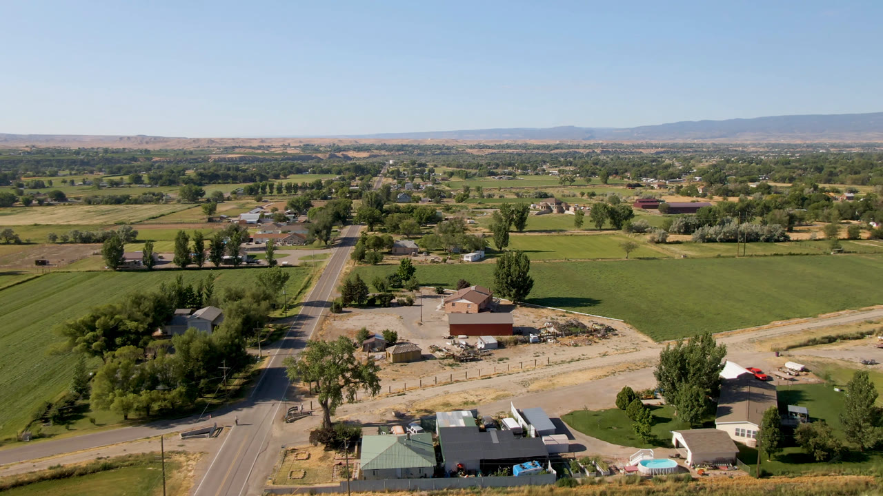 Aerial view in Palisades, Colorado with the road more centered, green crop fields on both sides, small homes and outbuildings below, distant mountains, and no visible people.
