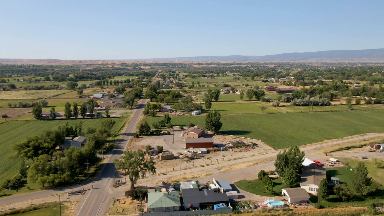Drone view of farmland, a road, clustered trees, outbuildings, and mountains under clear sky.