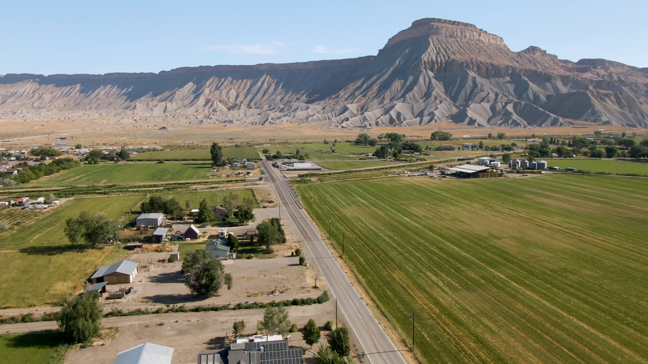 At 0:00, an aerial view in Palisades, Colorado shows green fields beside a straight road, scattered farm buildings, trees, and a broad mesa in the distance with no visible people.