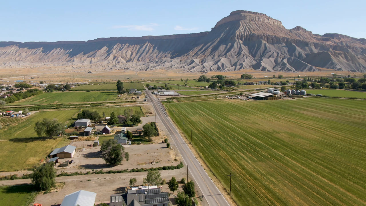 At 0:01, the drone view holds green agricultural rows, a paved road, small rural structures, and tan eroded slopes rising behind Palisades, Colorado, with no visible people.