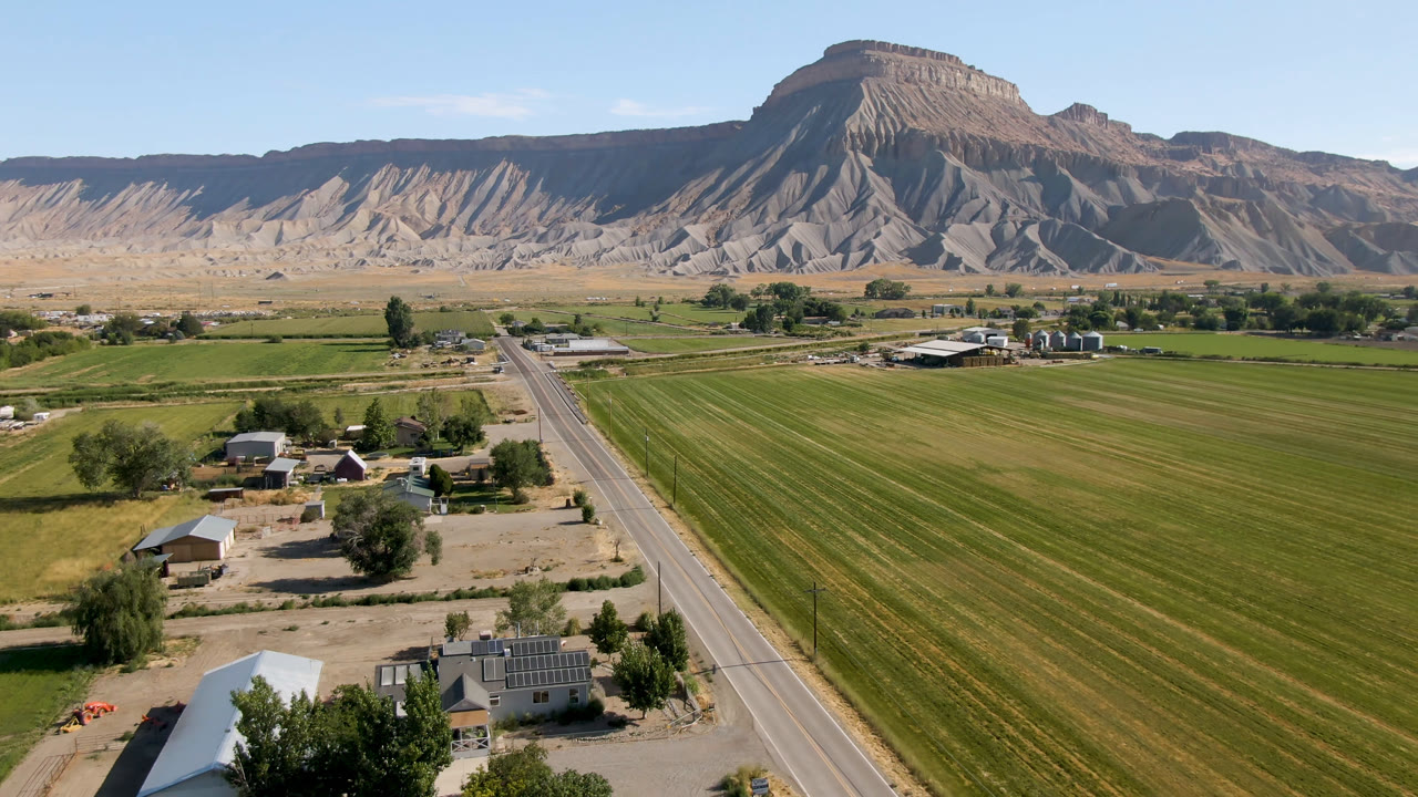 Aerial view in Palisades, Colorado with the road running through the lower frame, farm plots on both sides, low rural buildings, trees, and layered mesa cliffs beyond with no visible people.