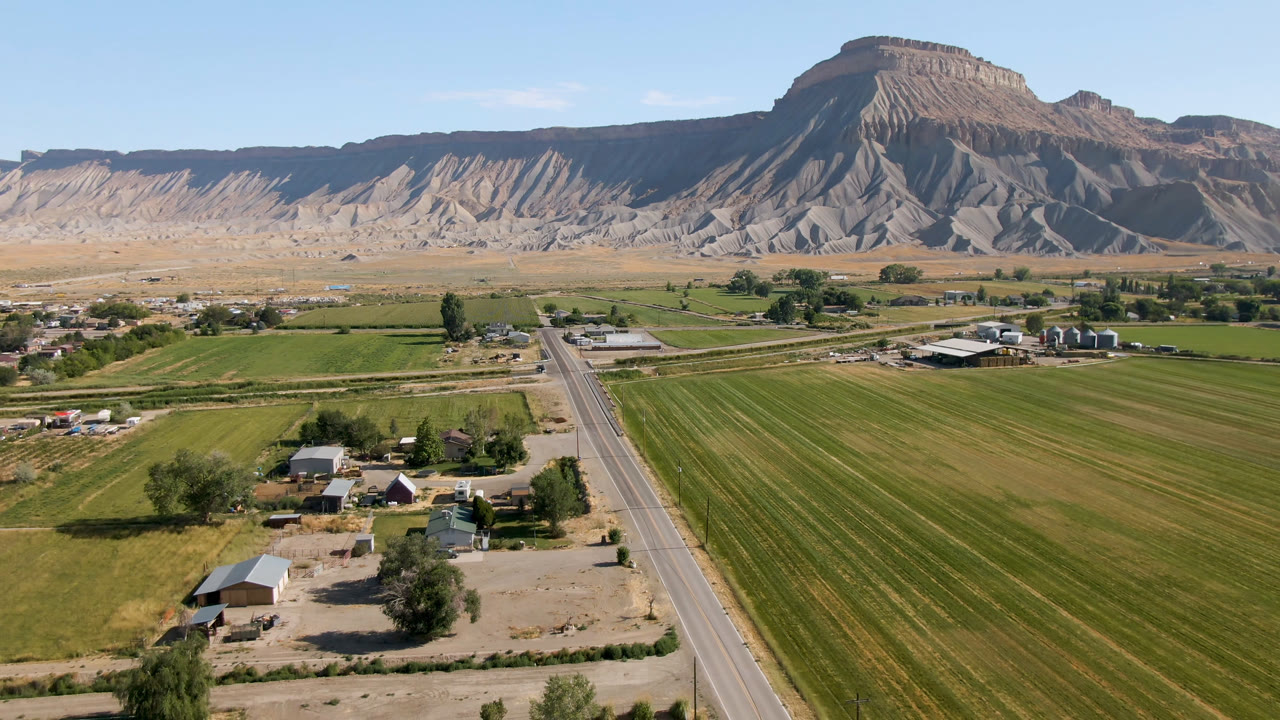 Second aerial frame over Palisades, Colorado, with the road running from the lower frame toward farms and fields below a flat-topped mountain formation, with no visible people.