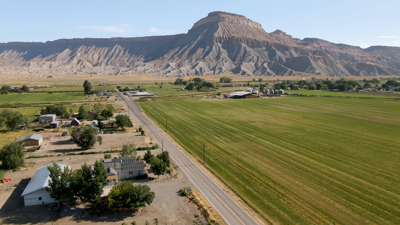 Drone-height view in Palisades, Colorado with a rural road dividing homes and green fields, low buildings near trees, mesa cliffs beyond, and no visible people.
