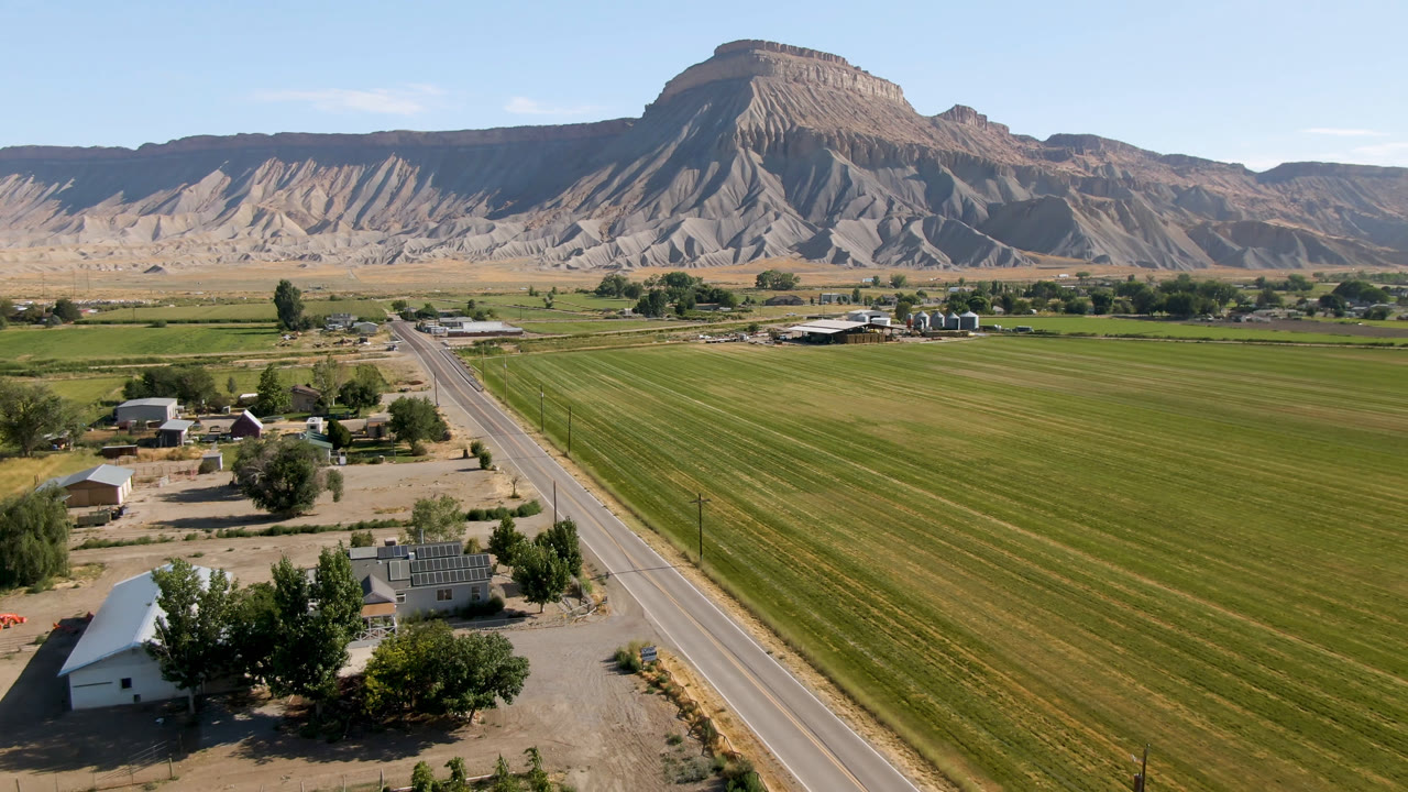Aerial farmland scene with road, house cluster, trees, fields, and distant mesa.