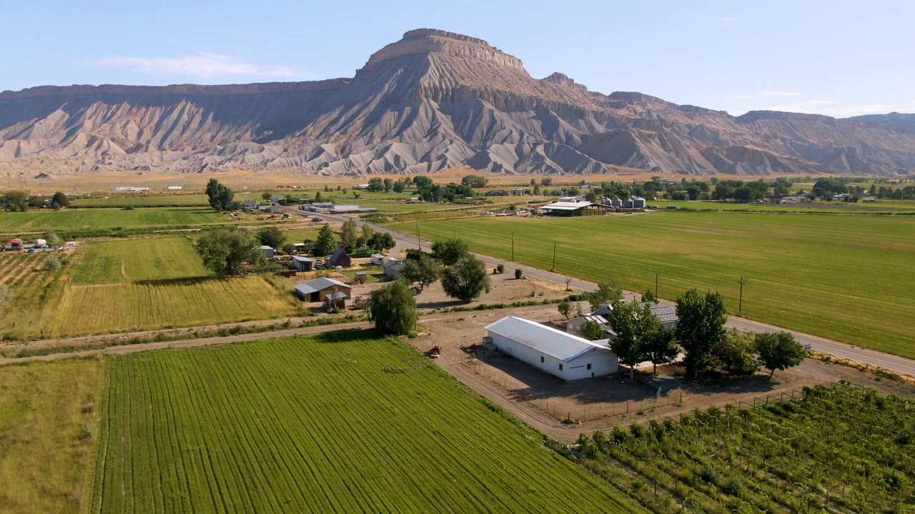 Opening frame of Palisades, Colorado farmland, with green fields in front, scattered farm buildings near a road, a tan mountain ridge in back, and no visible people.