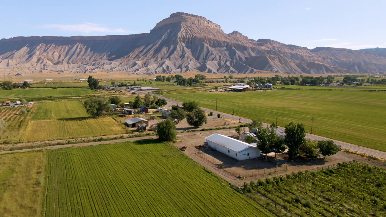 Third frame of the aerial view in Palisades, Colorado, with rectangular green fields leading to small rural buildings and a flat-topped mountain formation under clear sky, with no visible people.