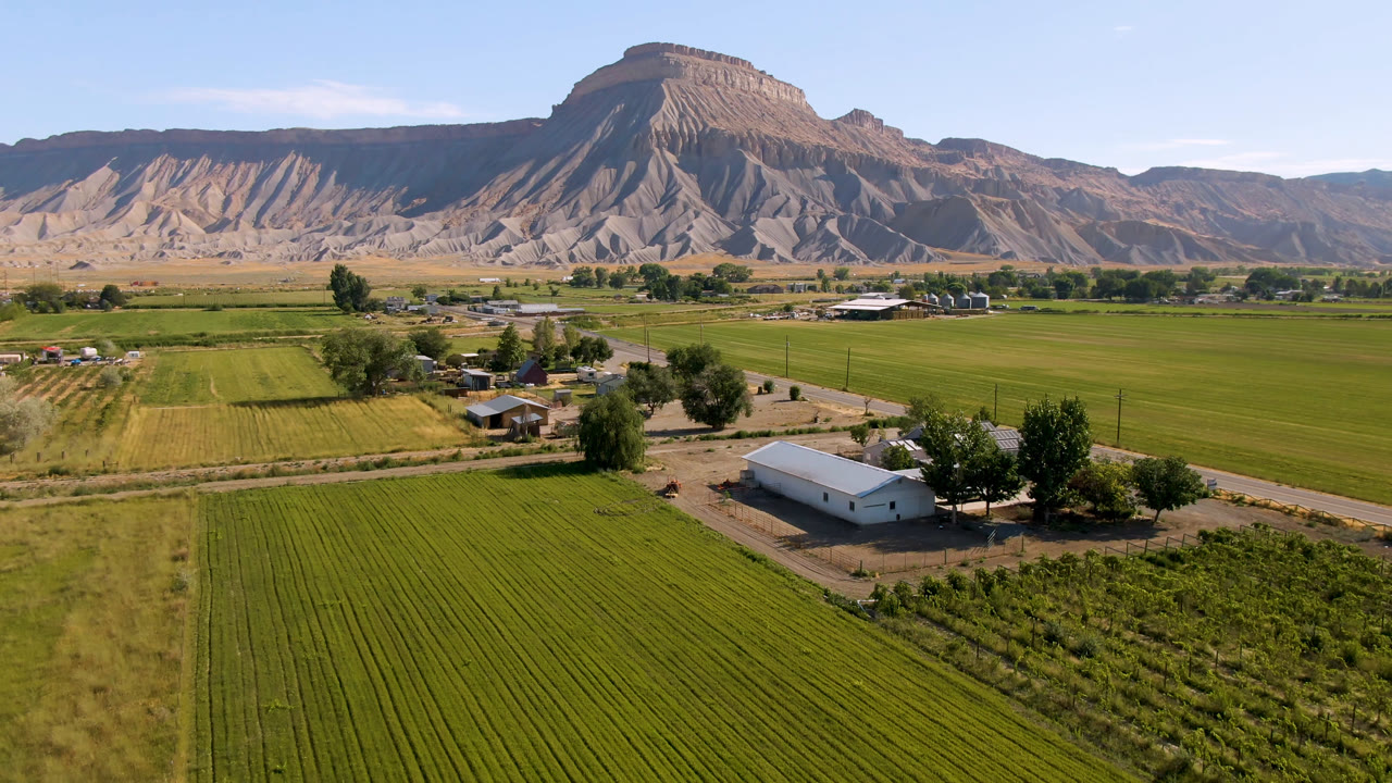 Second aerial frame in Palisades, Colorado showing the white farm building lower right of center, patterned green fields, roadside trees, dry open ground, the mountain ridge, and no visible people.