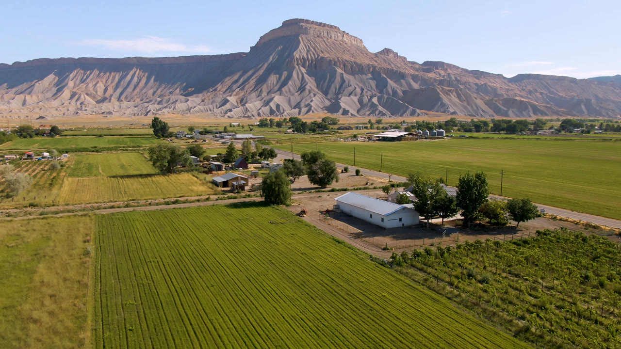 Third frame over Palisades, Colorado with rectangular green fields in the foreground, a cluster of rural structures and trees near the center, a long mountain ridge in back, and no visible people.
