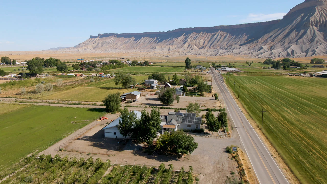 Wide aerial frame from Palisades, Colorado with a road dividing green farm fields, scattered houses and trees below, and ridged cliffs across the horizon, with no visible people.