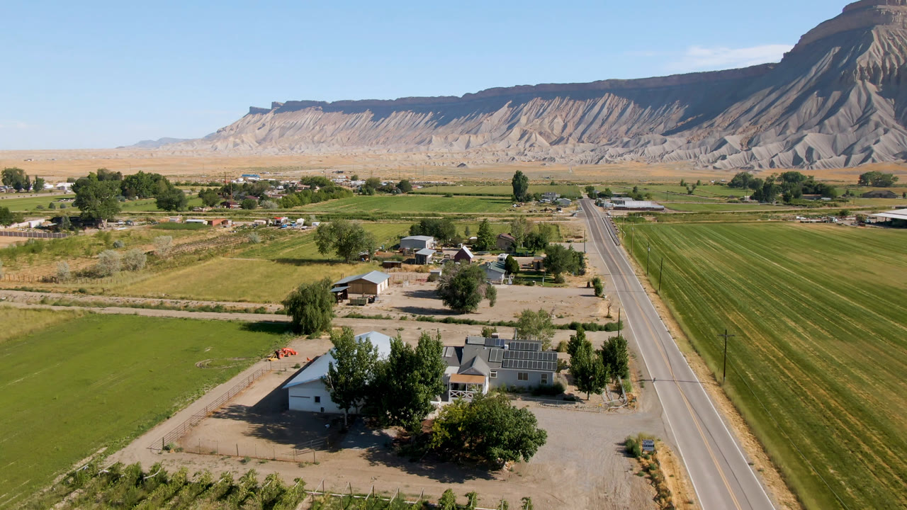 Rural road beside green fields and houses with a mesa behind them.
