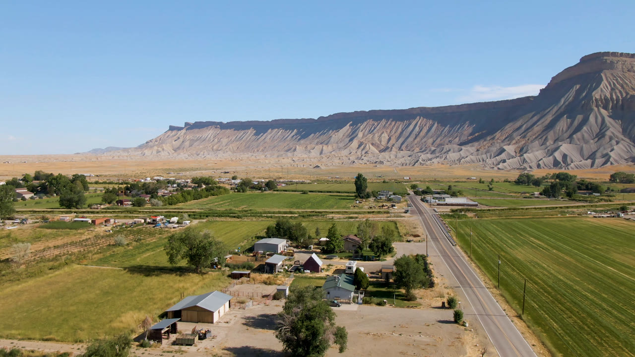 Opening aerial frame over Palisades, Colorado farmland with a straight road leading toward scattered homes and a mesa backdrop, with no visible people.