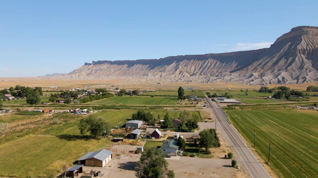 Aerial frame in Palisades, Colorado showing the road centered between green fields and homes, with pale mesa cliffs across the background and no visible people.
