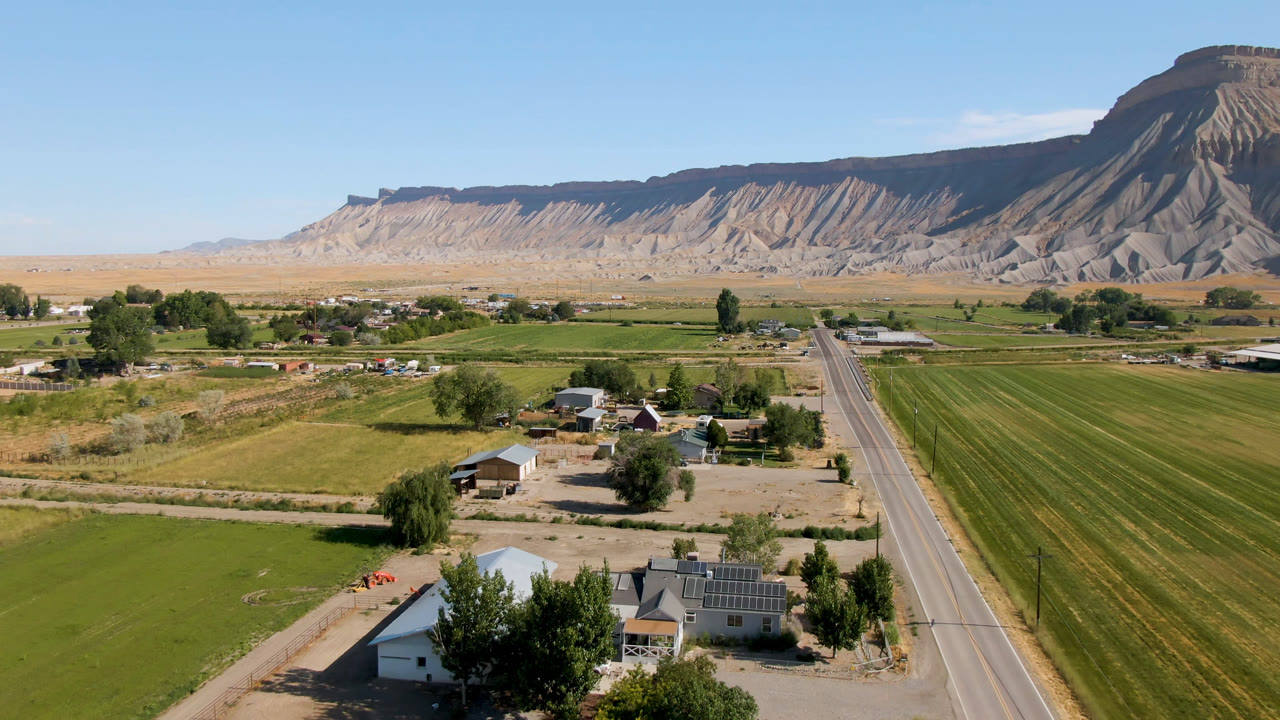 Aerial view of farm fields, a road, houses, and a mesa under clear sky.