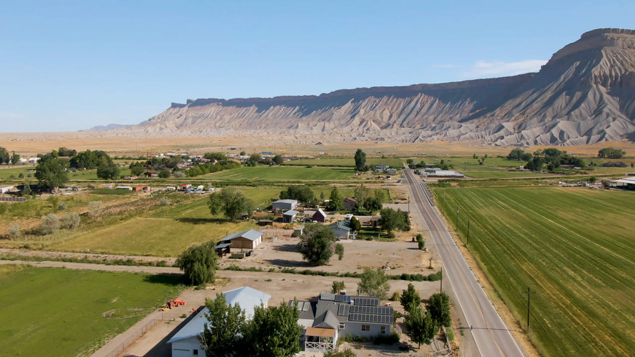 Aerial frame in Palisades, Colorado with the road nearer the center, green agricultural rows to the right, small buildings and trees below, and the mesa ridge spanning the background, with no visible people.