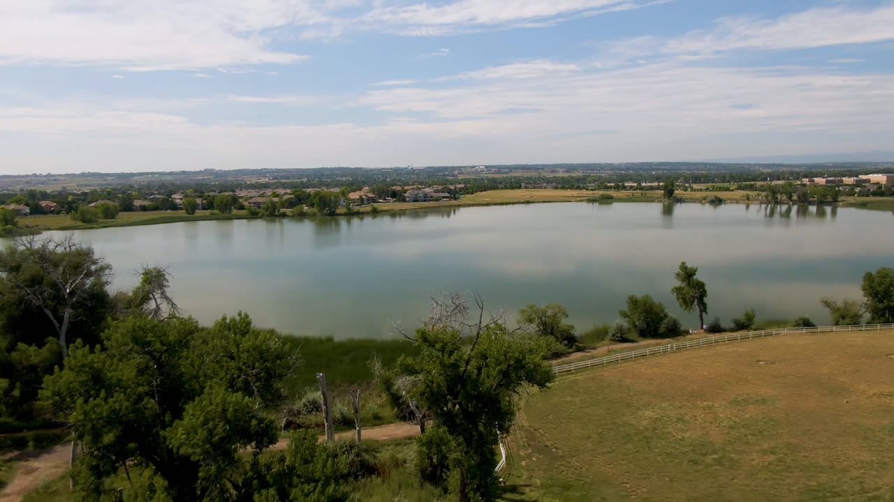 Wide aerial view over a large lake in Broomfield, Colorado, with trees and a fenced brown field in the foreground, distant low buildings across the water, and no visible people.