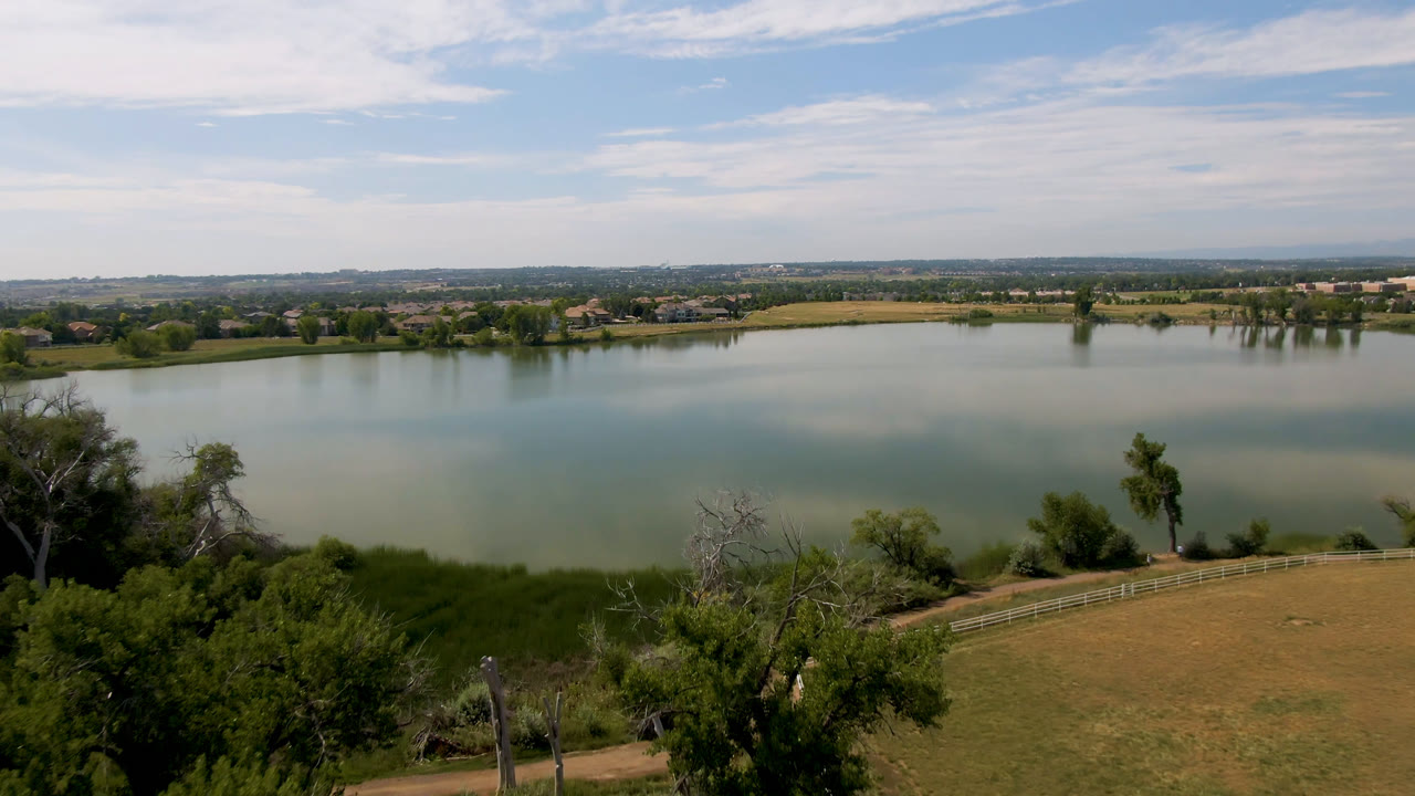 Elevated view of the Broomfield lake with green shoreline vegetation in front, calm water through the center, a distant shore with small buildings, and no visible people.