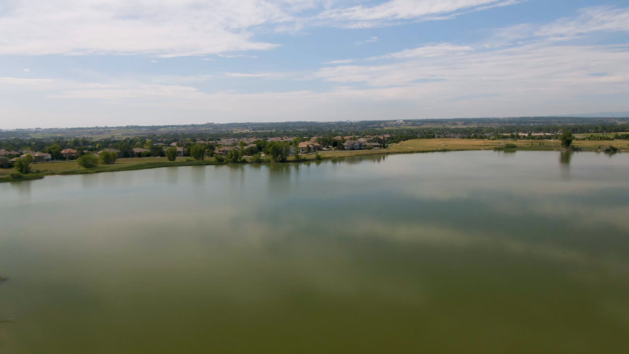 Opening frame of a wide Broomfield, Colorado lake view with green-blue water in the foreground, a low shoreline with trees and buildings in the distance, and no visible people.