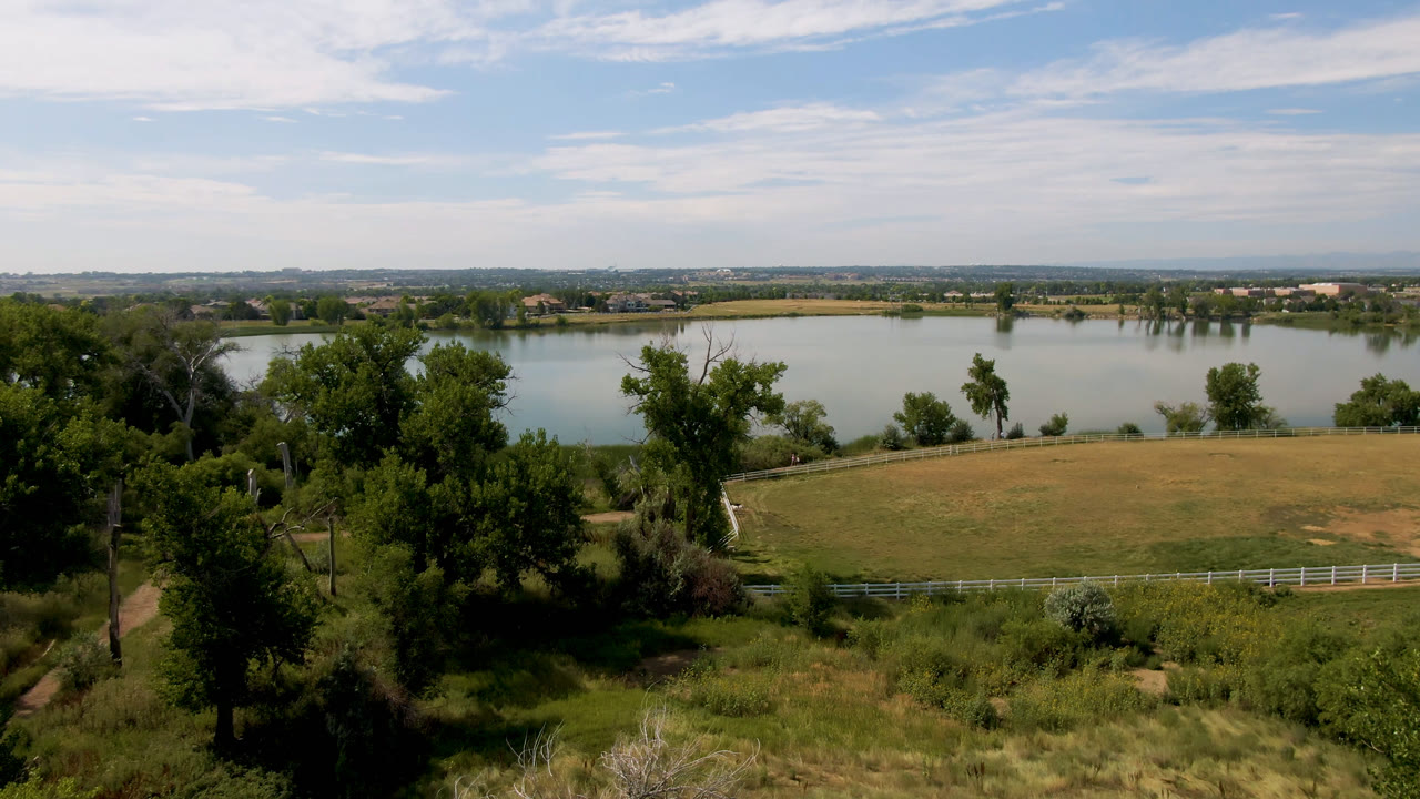 Opening view in Broomfield, Colorado, with a lake beyond foreground trees, grass, a white fence, and a tan field, with no visible people.