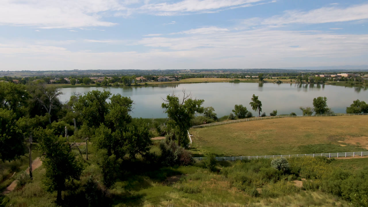 Broomfield, Colorado lakeside frame with dense foreground trees, a tan field to the right, calm water across the middle, and no visible people.