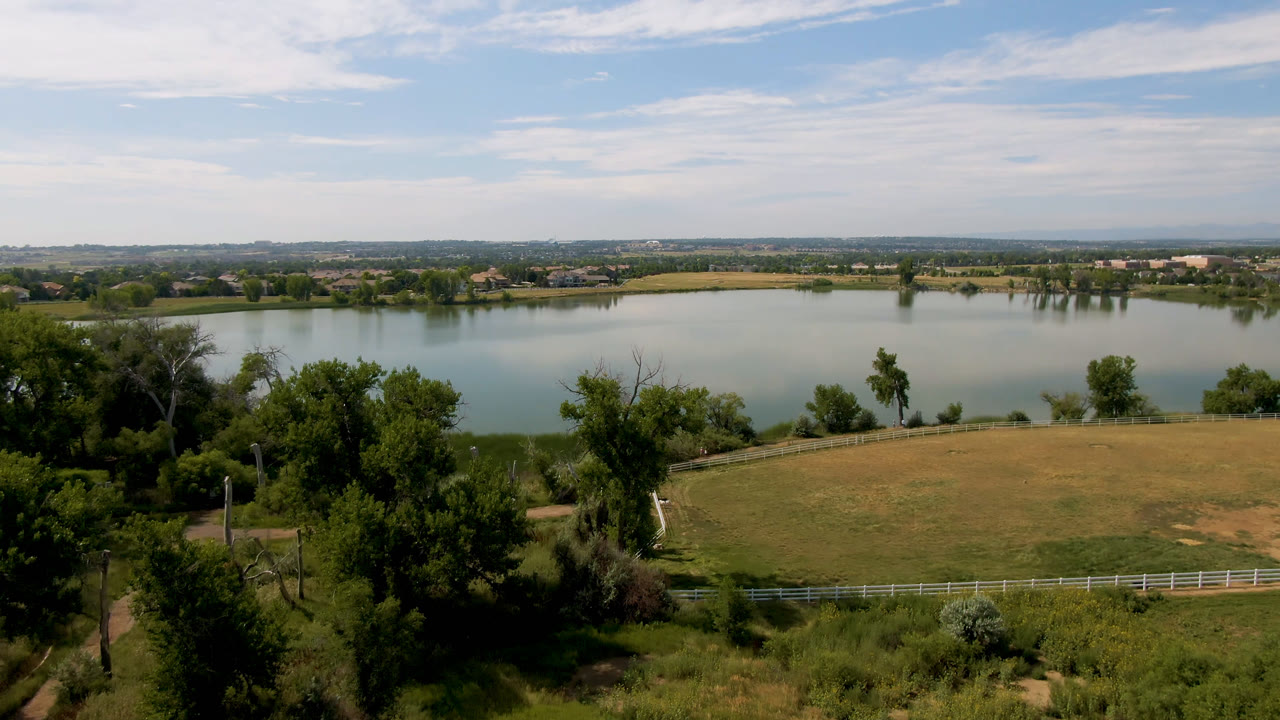 Calm lake with trees, grass, field, fence, and distant low buildings.