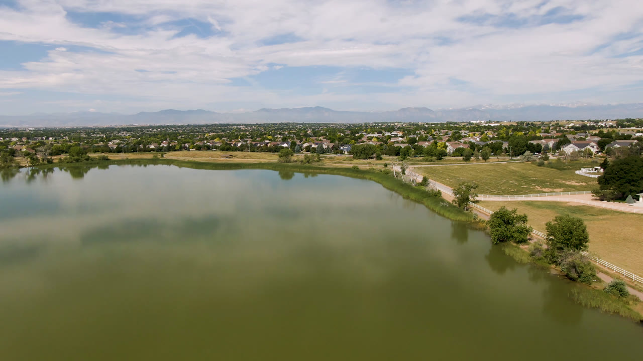Opening aerial frame over a green lake in Broomfield, Colorado, with a curving shoreline, dirt road, dry fields, trees, suburban buildings, distant mountains, scattered clouds, and no visible people.