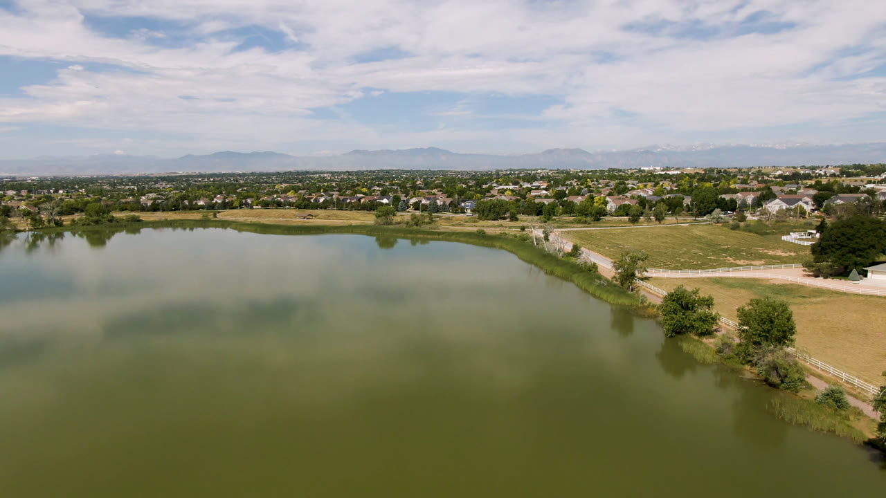 Second aerial frame in Broomfield, Colorado, showing broad green water in the foreground, a road tracing the shore, open fields, neighborhood buildings, trees, hazy mountains, and no visible people.