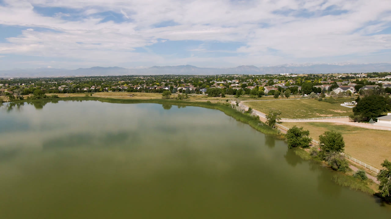Aerial view across a lake toward fields, trees, suburban buildings, and a mountain horizon.