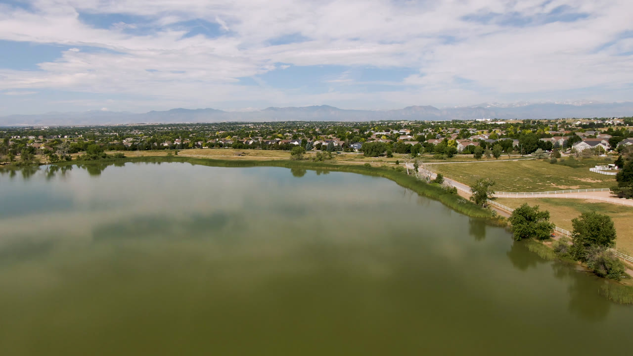 Aerial view of a green lake with shoreline trees, distant homes, mountains, and clouds.