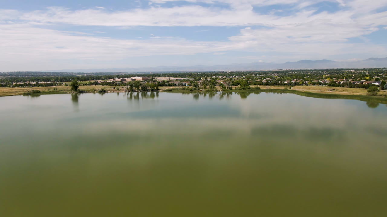 Wide aerial view over a green lake with fields and distant homes under a partly cloudy sky.