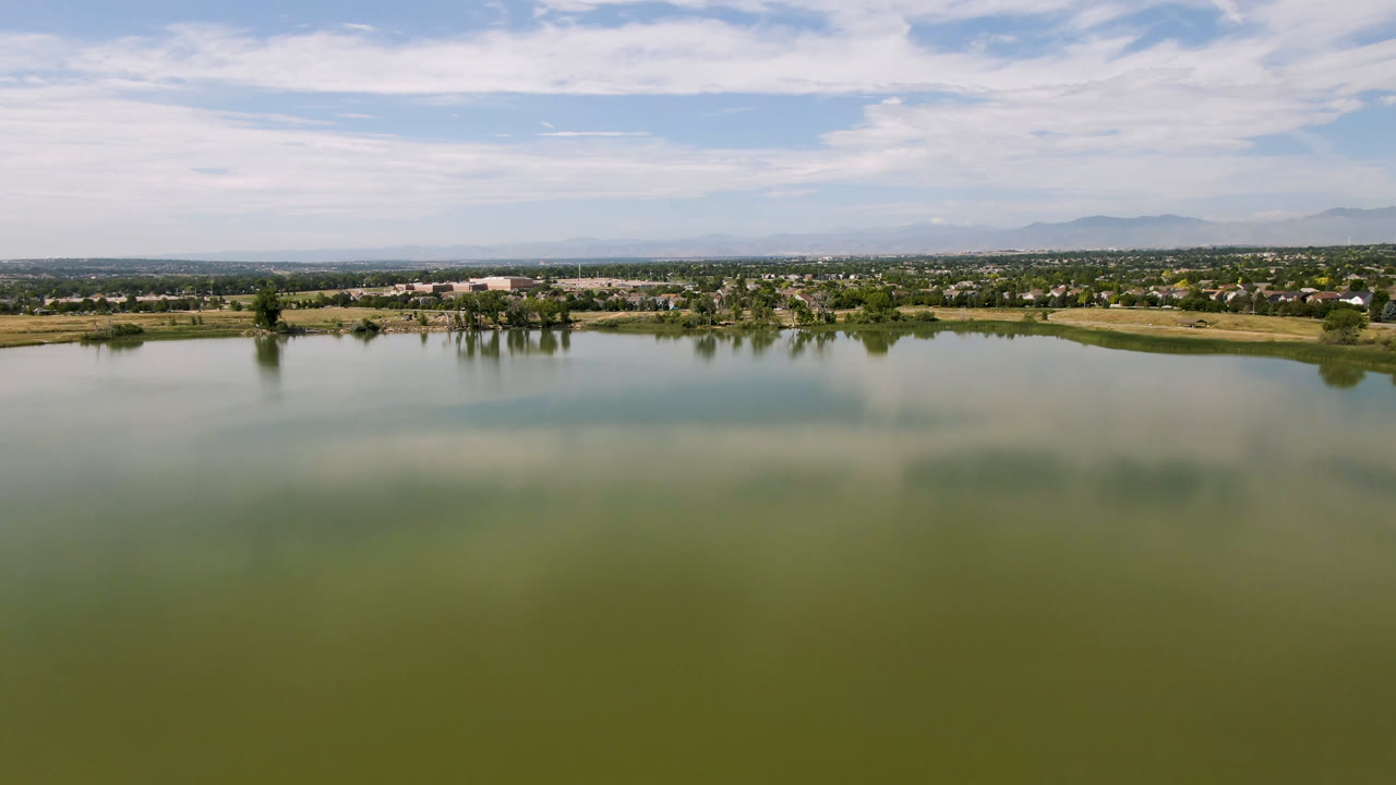 At 0:01, the Broomfield lake remains centered with broad green water below, a compact island or protruding shoreline in the midground, distant development along the horizon, and no visible people.