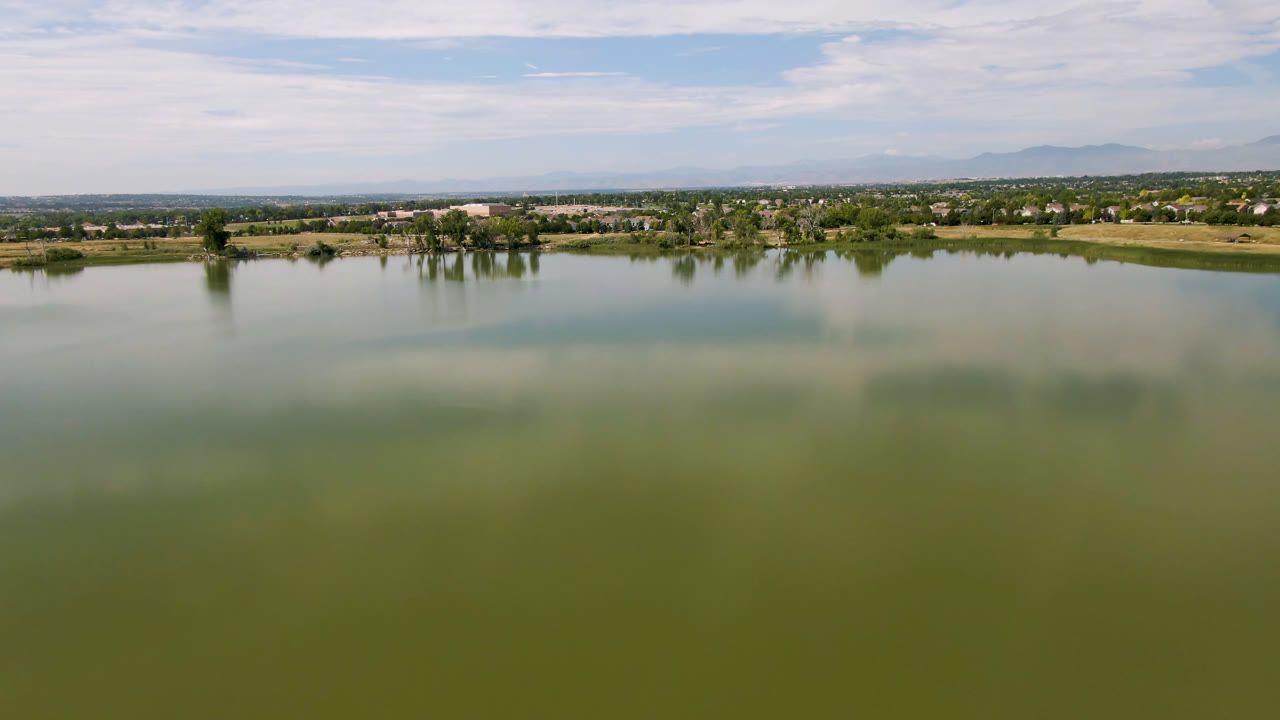 Wide opening view of a Broomfield, Colorado lake with green-blue water in the foreground, a tree-lined far shore, low buildings, distant mountains, and no visible people.