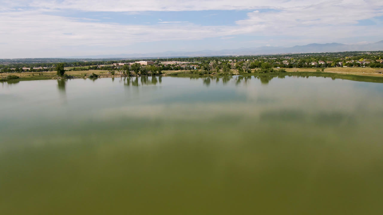 Second view of the Broomfield lake showing a broad muted-green water surface, shoreline trees and buildings across the center, hazy mountains, streaked clouds, and no visible people.
