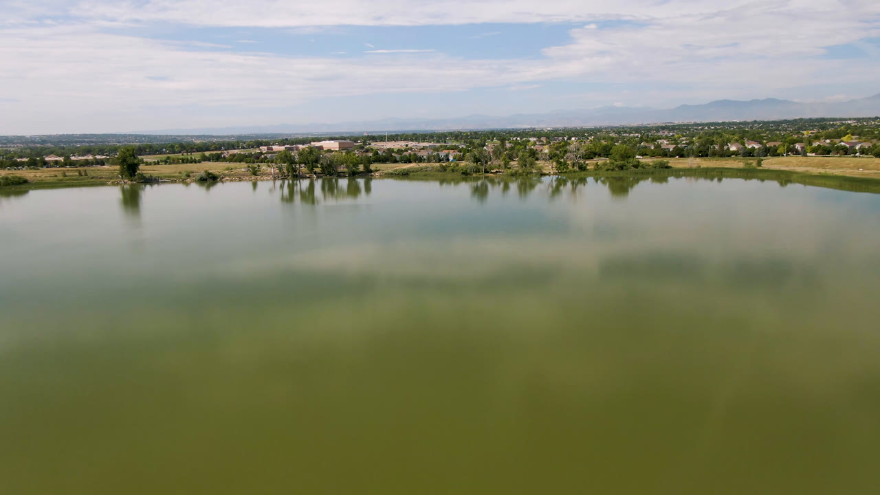 Lake foreground with distant trees, buildings, open land, mountains, and bright cloud cover.