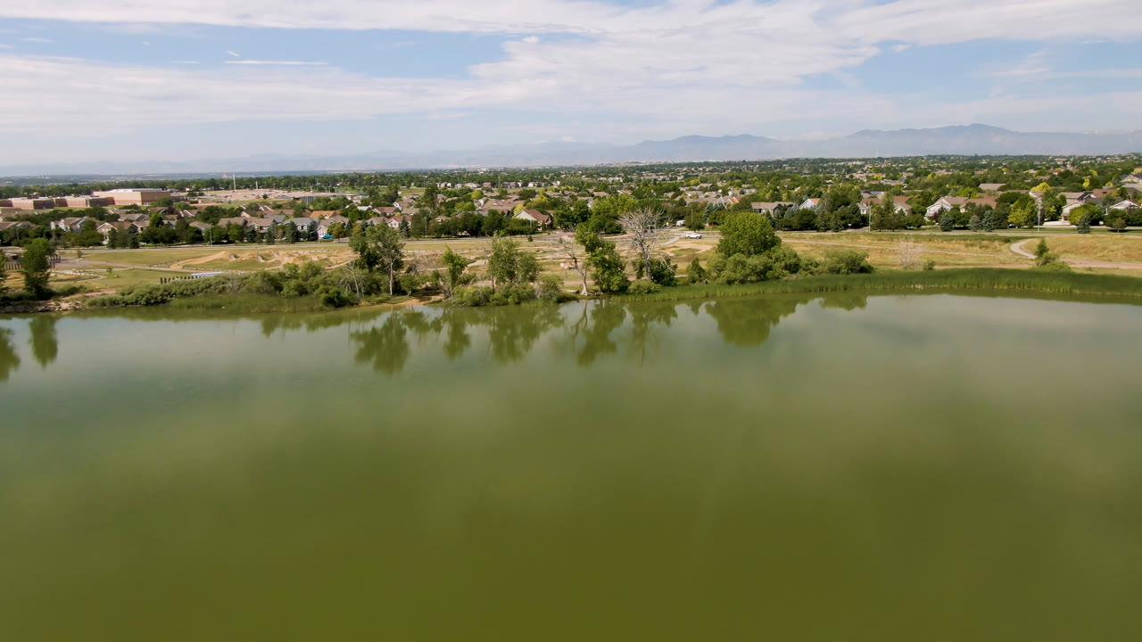 Aerial frame in Broomfield, Colorado with green lake water below, a tree-lined shore, open grass fields, rows of distant homes, a hazy mountain horizon, and no visible people.