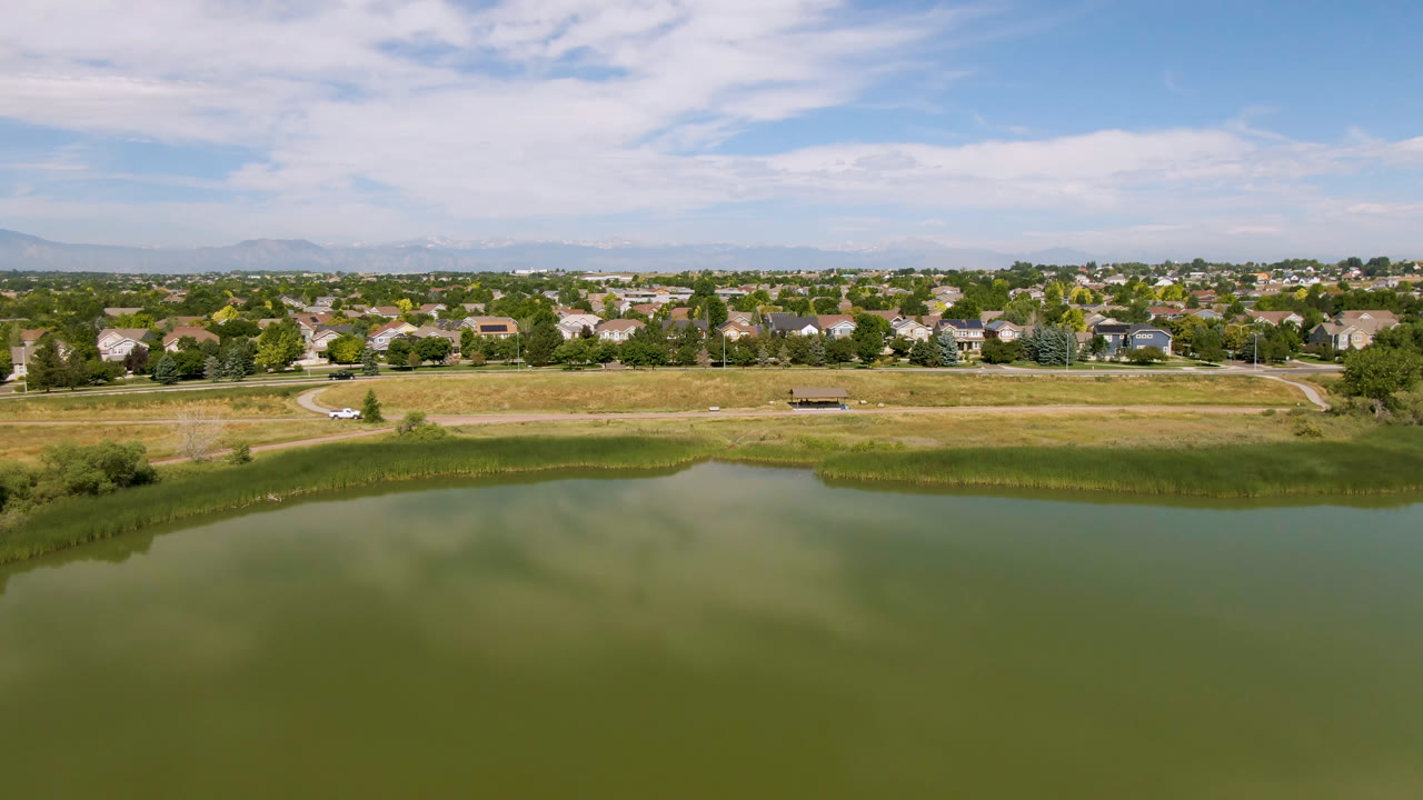 Wide aerial view of green water, grassy shoreline, suburban homes, and cloudy blue sky.