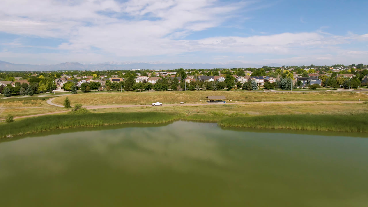 Wide aerial frame in Broomfield, Colorado showing green lake water in front, reeds and dry grass beyond it, a road, suburban homes, distant mountains, and no visible people.