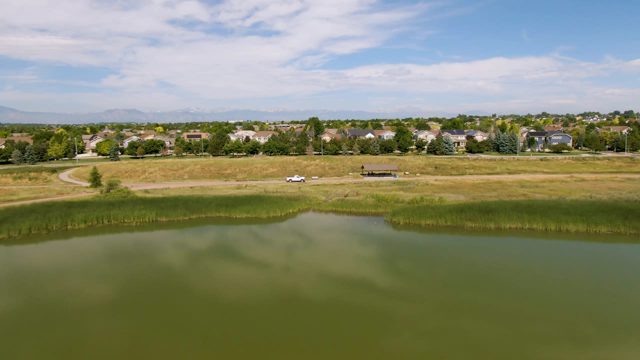 Broomfield, Colorado aerial view with green water and cloud reflections below, tall reeds along the shore, a dry field, a white vehicle on the road, homes on the horizon, and no visible people.