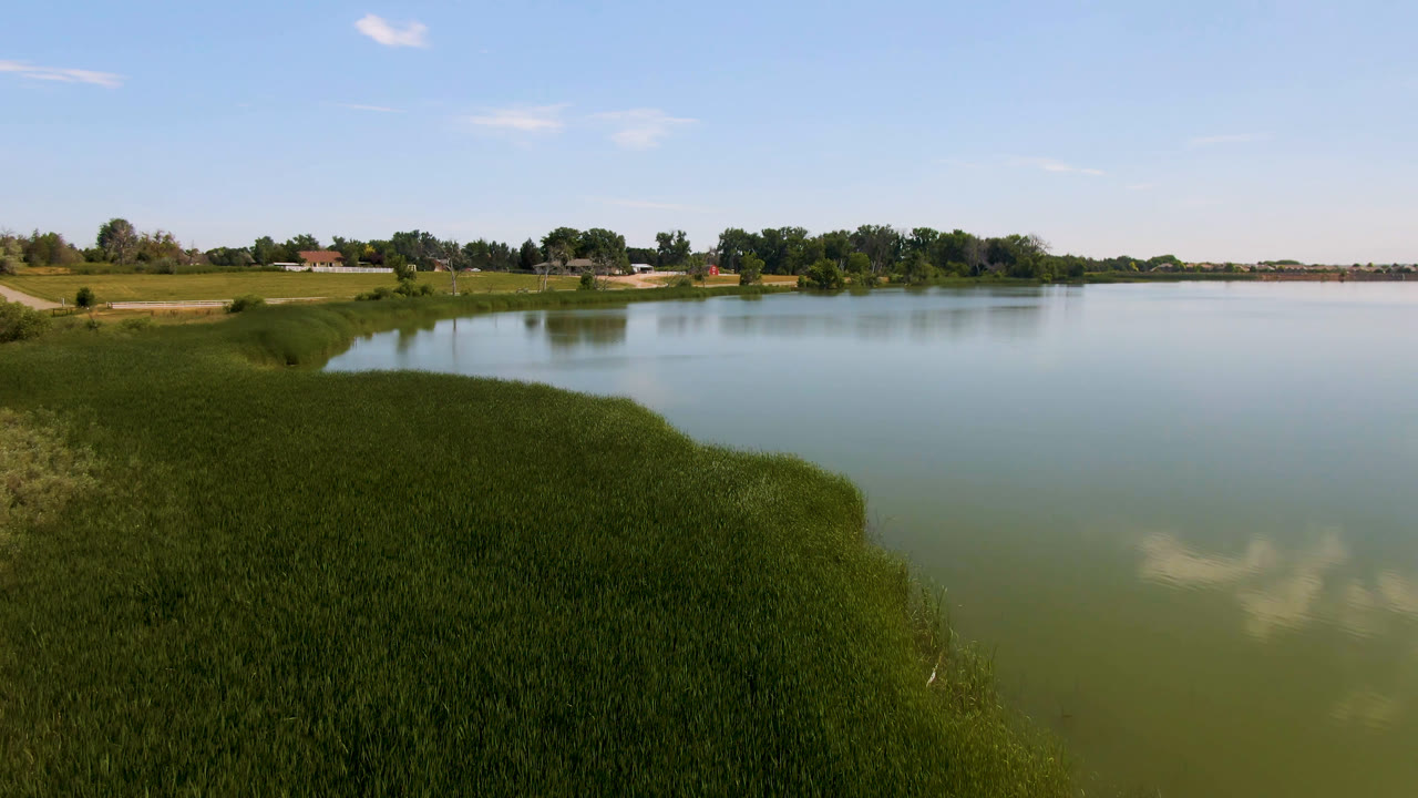 Second view of the Broomfield lake shows foreground reeds curving around the water, a pale sky reflection, distant trees, small buildings, and no visible people.