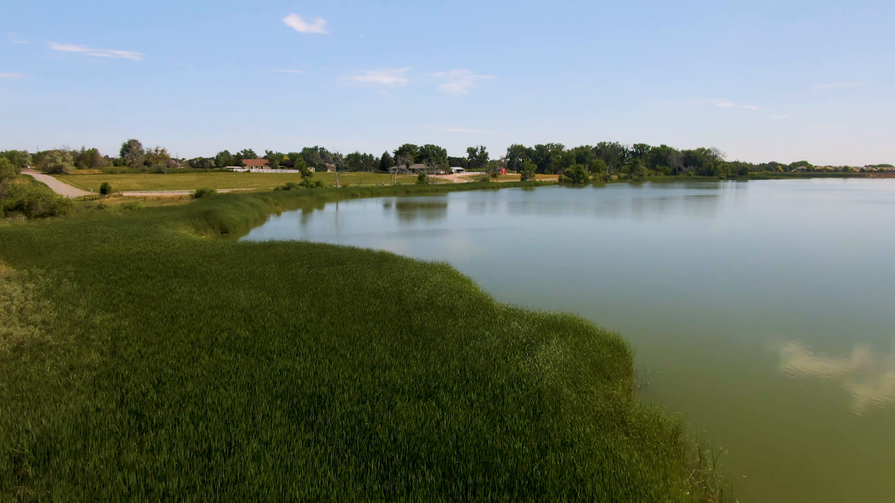 At one second, the Broomfield lake remains framed by thick foreground grass, broad pale water, a far tree line, small distant buildings, and no visible people.