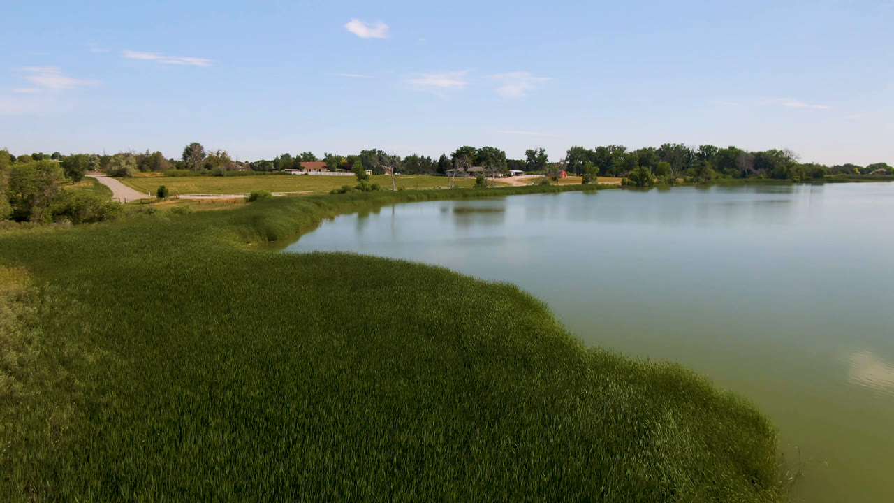 Broomfield, Colorado lake view with the curved grassy bank in the foreground, still water across the center, far trees and low buildings, and no visible people.