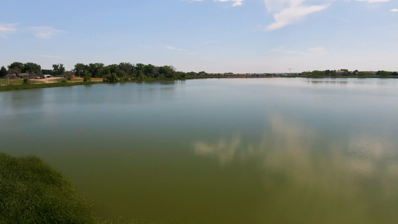 Wide lake with grassy bank, trees, distant buildings, blue sky, and cloud reflections.