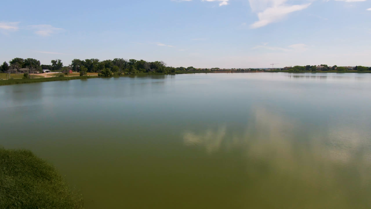 Broomfield, Colorado lake scene with open green-blue water, a low tree line, small distant houses, a bright sky, cloud reflections, and no visible people.