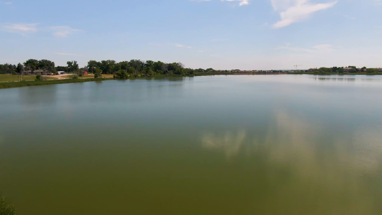 Wide lake with distant trees, grassy bank, blue sky, and cloud reflections.